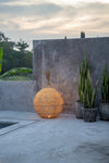Woven spherical lantern on a concrete surface with plants and a stone wall in the background.