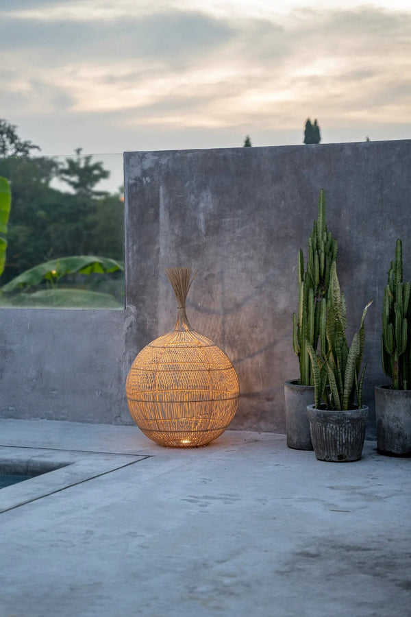 Woven spherical lantern on a concrete surface with plants and a stone wall in the background.