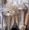 Two silver textured vases with white flowers and candles on a reflective surface.