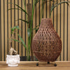 Woven rattan lamp on a wooden surface with plants and a striped wall in the background
