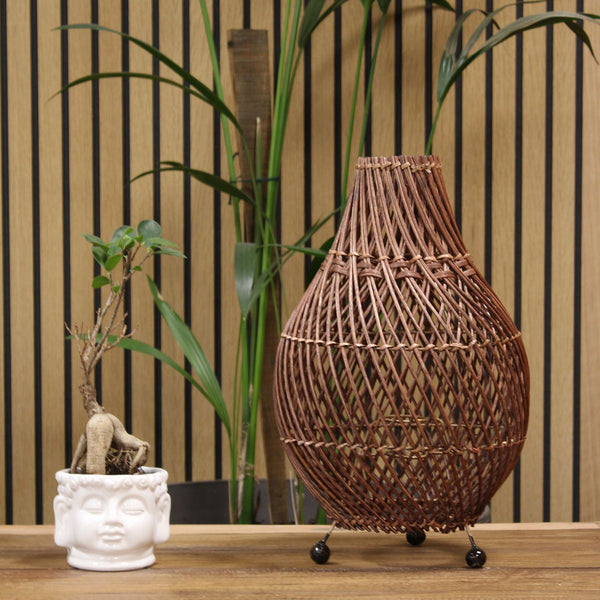 Woven rattan lamp on a wooden surface with plants and a striped wall in the background