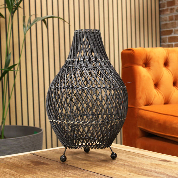 Woven rattan table lamp on a wooden surface with an orange sofa and plant in the background