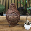 Woven brown lamp on a wooden surface with a plant in the background
