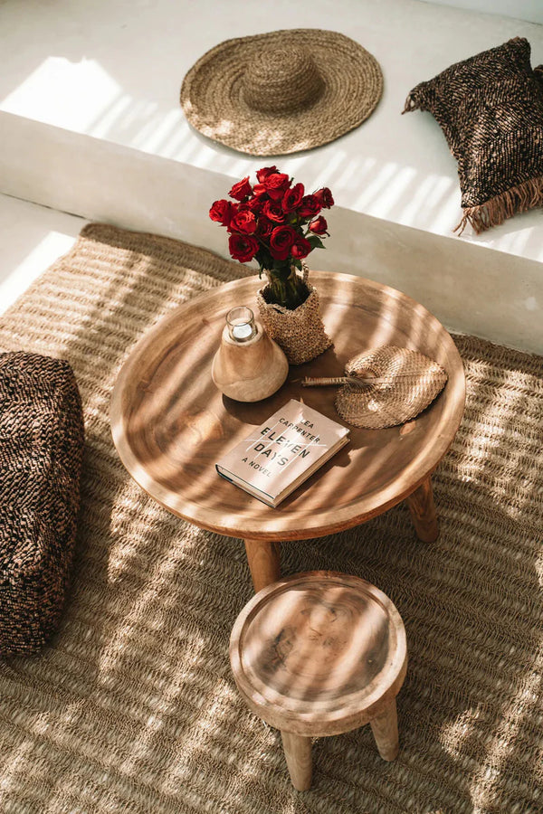Round wooden table with a vase of red flowers, a book, and a small stool on a textured floor.