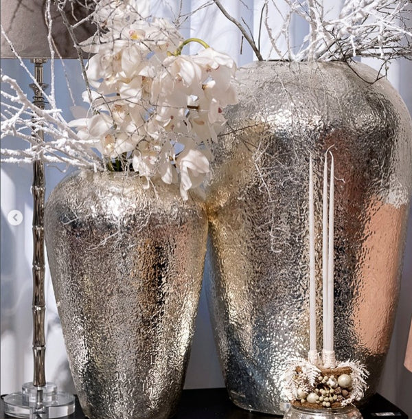 Two silver textured vases with white flowers and candles on a reflective surface.